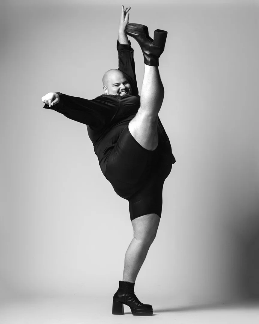 Full-body portrait of a bald adult performing a leg extension wearing a dark suit and platform boots against a neutral backdrop.