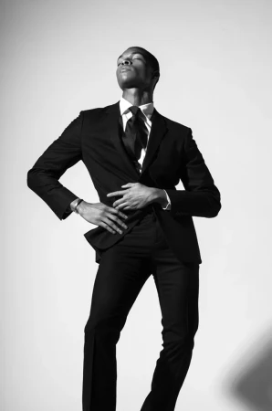 Black and white portrait of a Black male model wearing a suit with hands in front of his stomach in a light grey photo studio.