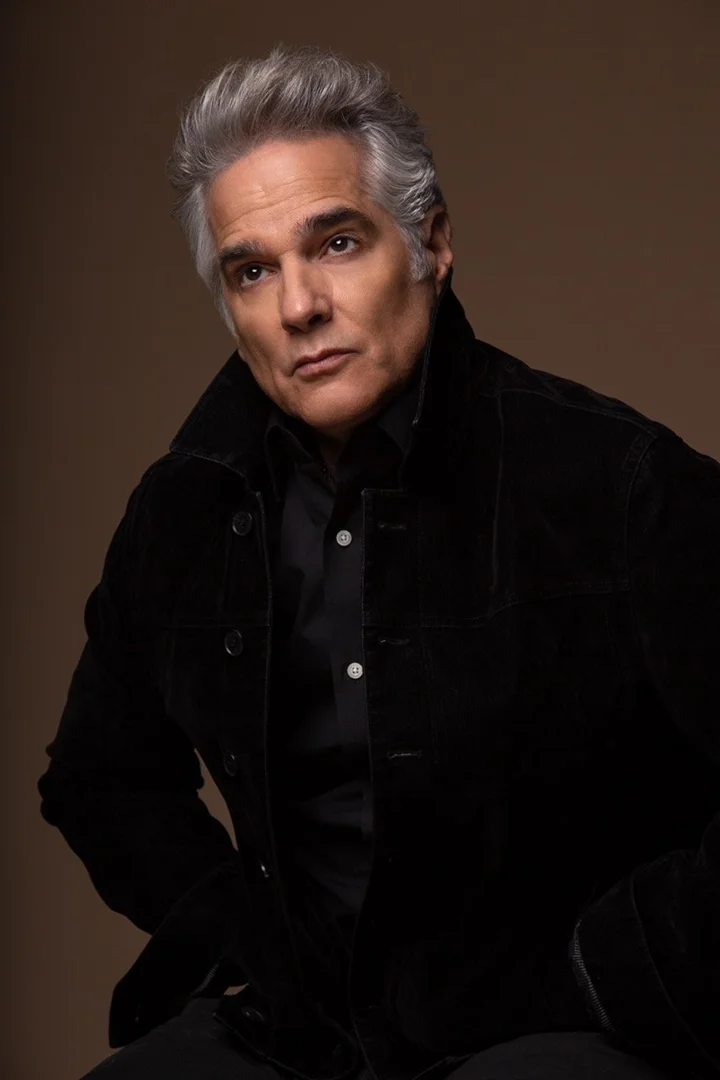 Portrait of an older man with grey hair wearing a black jacket against a dark background
