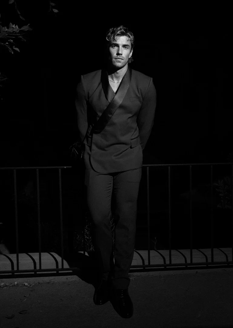 Black-and-white portrait of a handsome adult man in a dark suit with hands behind his back, standing outdoors at nighttime.