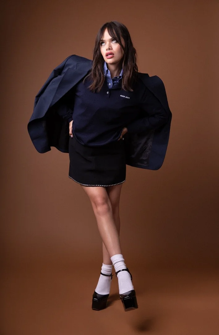 A young brunette woman poses in a blue top, black skirt, white socks, and black Mary Janes in a fashion studio shoot.