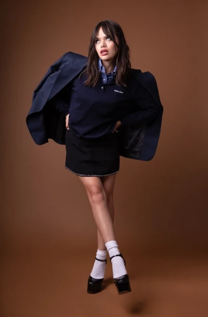 A young brunette woman poses in a blue top, black skirt, white socks, and black Mary Janes in a fashion studio shoot.