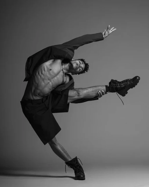 A Black male dancer with a muscular build is captured in a black-and-white studio shot, wearing combat boots and a partially open black jacket.