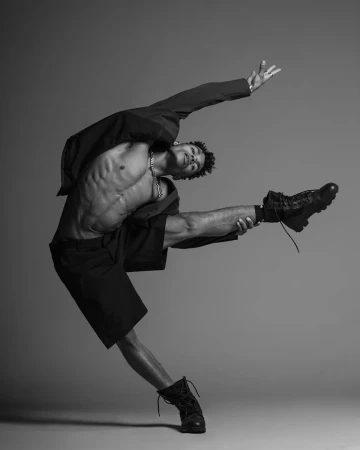 A Black male dancer with a muscular build is captured in a black-and-white studio shot, wearing combat boots and a partially open black jacket.
