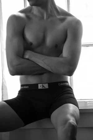 Black and white close-up of a muscular, shirtless adult male sitting near a window wearing branded underwear, indoors.