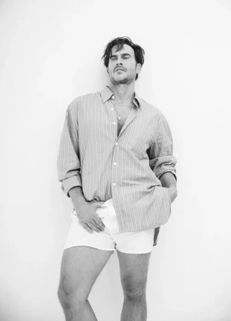 Man wearing a striped button-down shirt and white shorts standing against a seamless white studio background