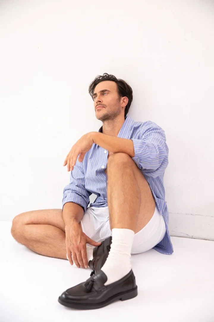 Man in blue striped shirt and white shorts sits against a white wall with one black loafer and white socks, looking off to the side.