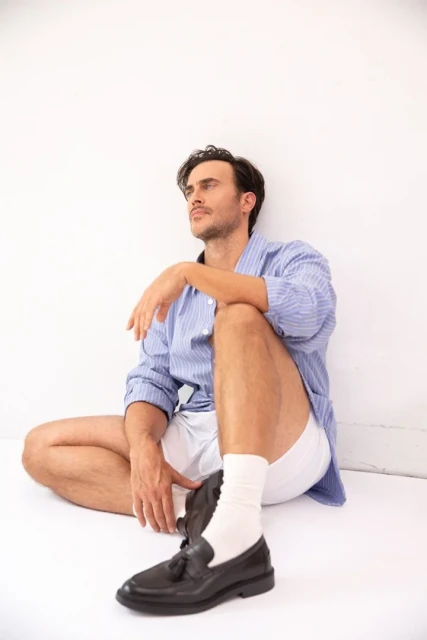 Man in blue striped shirt and white shorts sits against a white wall with one black loafer and white socks, looking off to the side.