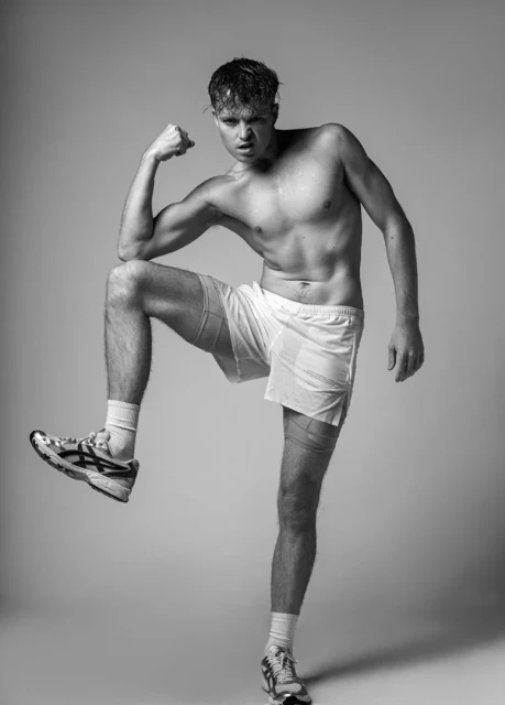 Athletic man poses shirtless in white shorts and sneakers in a studio. He's flexing and has a determined facial expression.