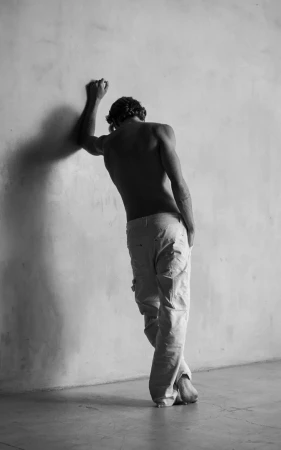 Black and white portrait of a slender, muscular adult male leaning against a textured wall in a photo studio, wearing white pants.