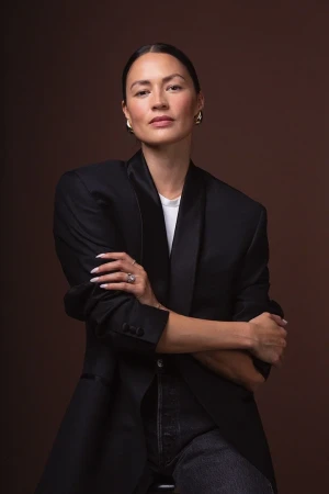 Portrait of a woman with dark hair in an oversized black blazer and white t-shirt against a brown background