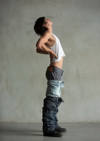 Man wearing a white tank top and multiple layers of denim jeans pulled down, arching back against a plain grey concrete wall.
