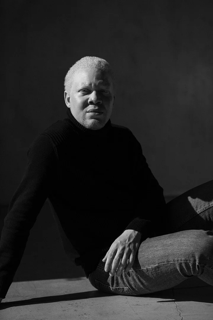 Monochrome portrait of an albino man in a black turtleneck sweater seated against a plain dark wall.