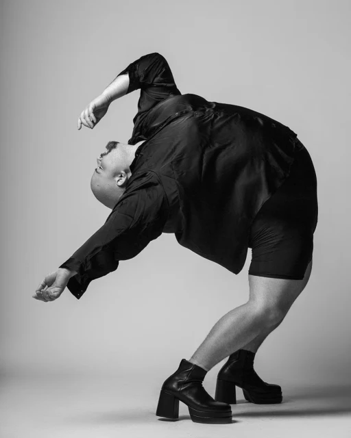 Monochrome portrait of bald person in a black shirt, shorts, and heeled boots, arching backwards against a grey backdrop in a studio.