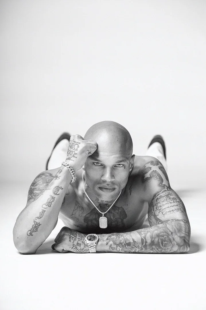 Monochrome portrait of a muscular, bald, and tattooed man lying on a white surface, striking a pose and looking intensely at the camera.
