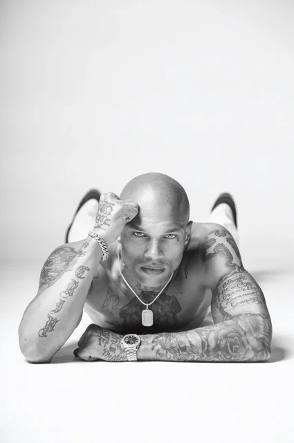 Monochrome portrait of a muscular, bald, and tattooed man lying on a white surface, striking a pose and looking intensely at the camera.