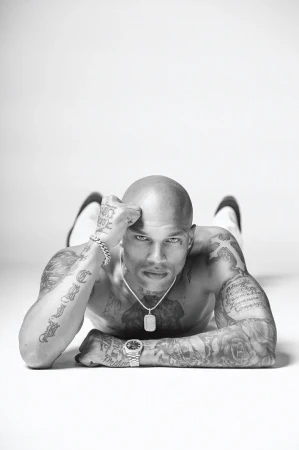Monochrome portrait of a muscular, bald, and tattooed man lying on a white surface, striking a pose and looking intensely at the camera.