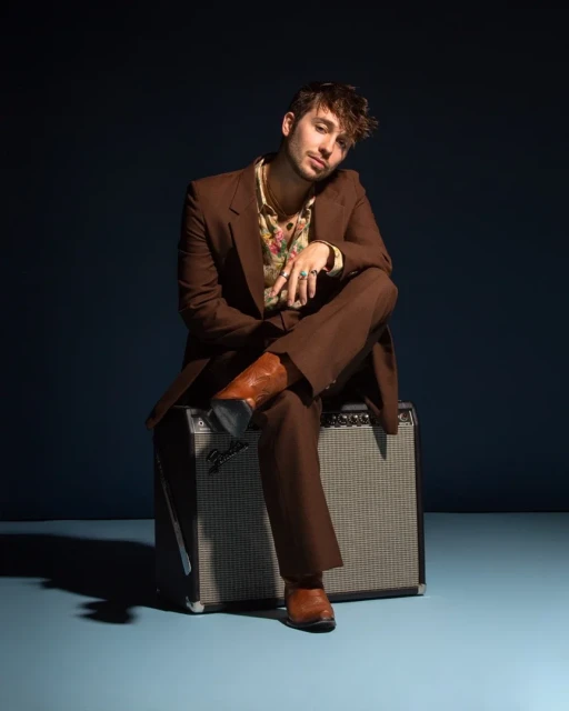 Portrait of a young adult male in brown suit seated on a vintage amplifier in a studio with cool background and warm lighting.