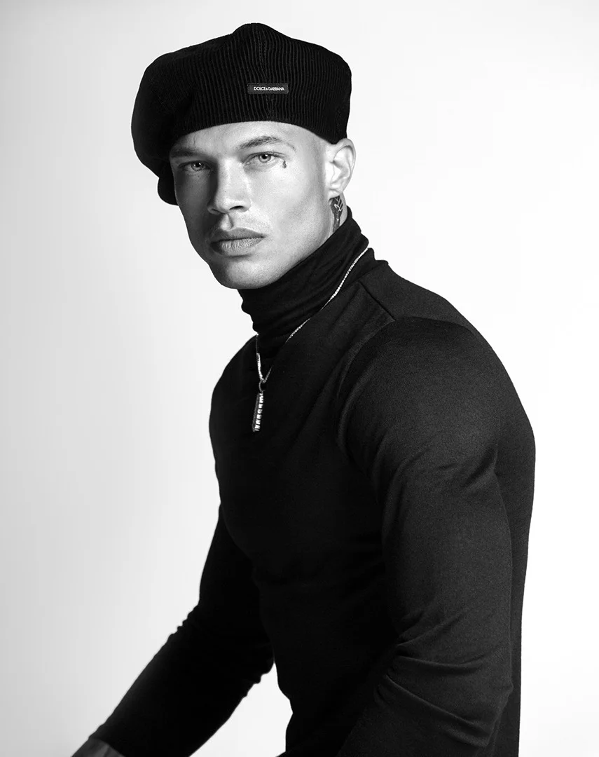Black and white portrait of an adult male fashion model wearing a beret, turtleneck shirt, and jewelry against a white backdrop.