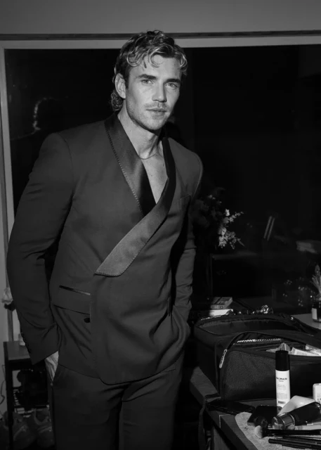 Black and white portrait of a blond adult male in a suit, posed indoors in a low-light environment with visible hair and makeup products.