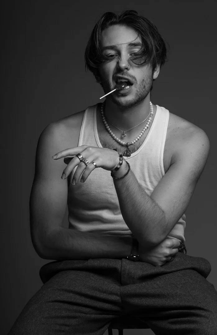 Monochrome portrait of a young man wearing a white tank top and pearl necklace while holding a lollipop in his mouth.