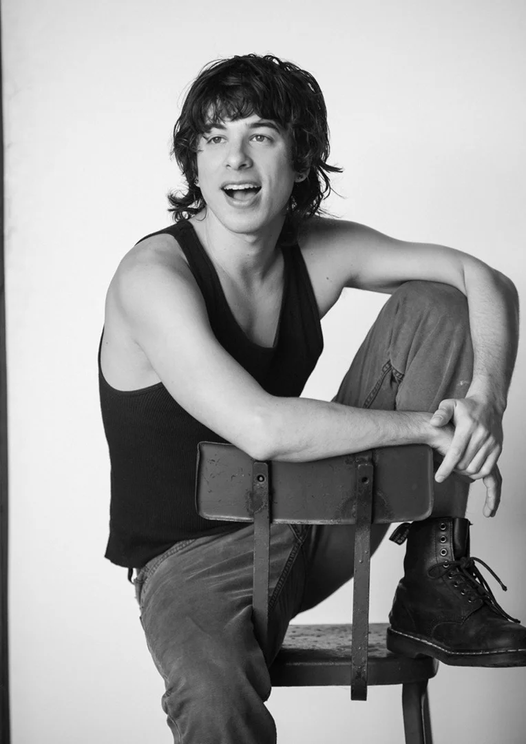 Monochrome portrait of a young adult male sitting on a chair, wearing a tank top and jeans, gazing to the side in a photo studio setting.