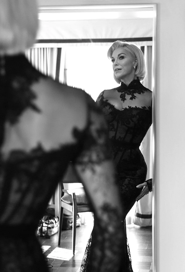 Black and white photo of a woman in a black lace gown, reflected in a mirror, posing in an indoor setting, portrait orientation.