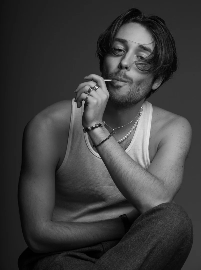 Monochrome portrait of a young man wearing a ribbed white tank top, pearl necklace, and rings, holding a lollipop.