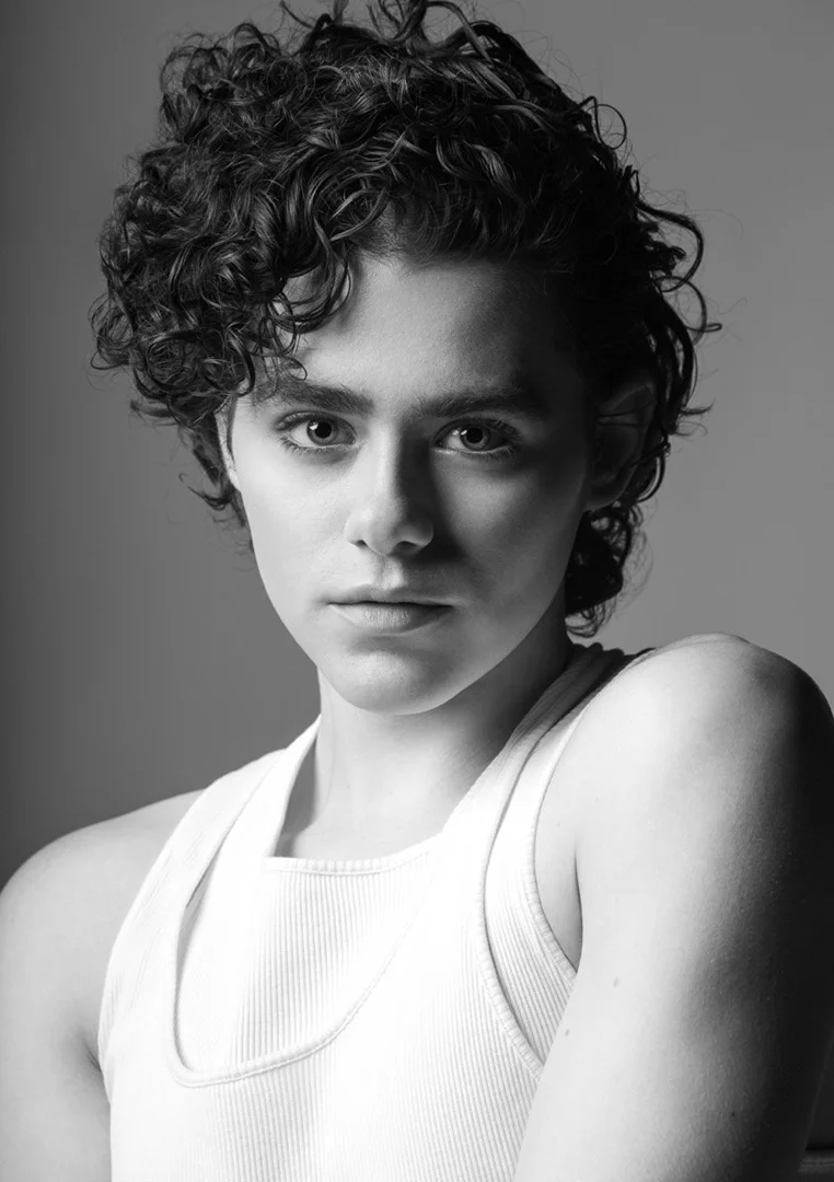 Black and white portrait of a young man with curly hair wearing a white ribbed tank top against a grey studio backdrop.