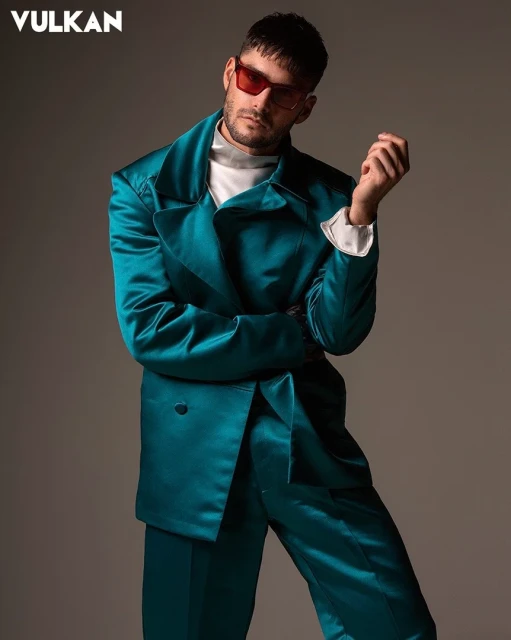 Stylish man in teal suit and sunglasses poses in studio. Vulkan Magazine logo in corner. Portrait orientation, fashion editorial image.