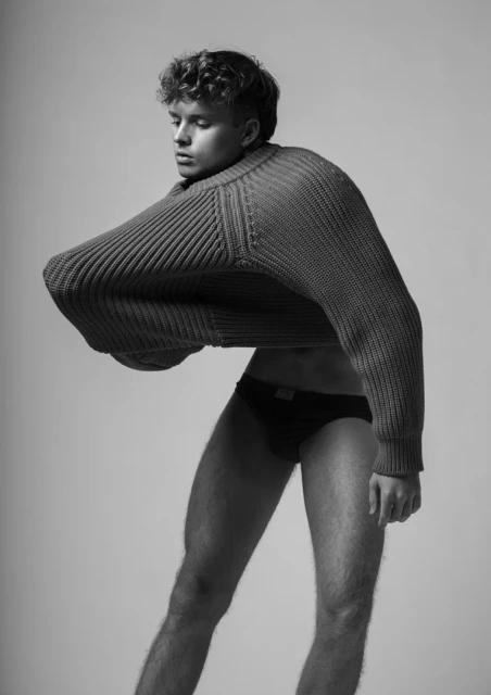 Monochrome fashion portrait of a male model with curly hair wearing a large knit sweater and posing in underwear on a neutral backdrop.
