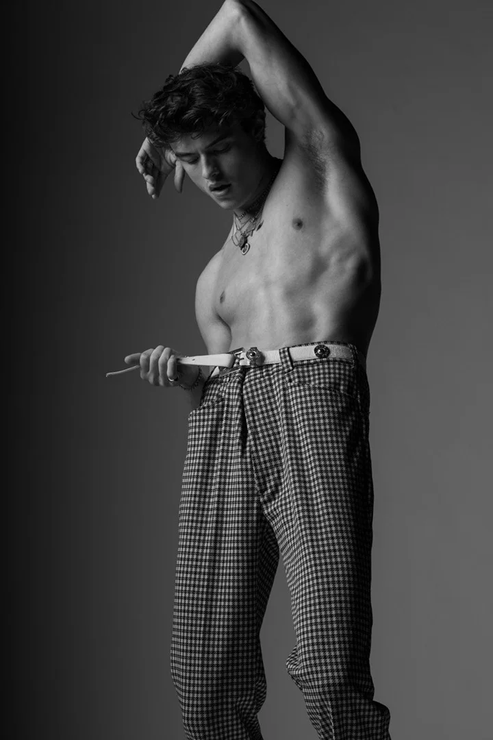 Shirtless young man wearing patterned trousers and a light belt, posing with arms raised in studio portrait