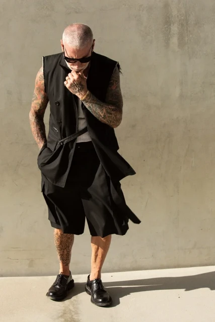 Mature man with bald head and tattoos wears dark vest and shorts, posing outdoors against a neutral concrete wall, looking downward.