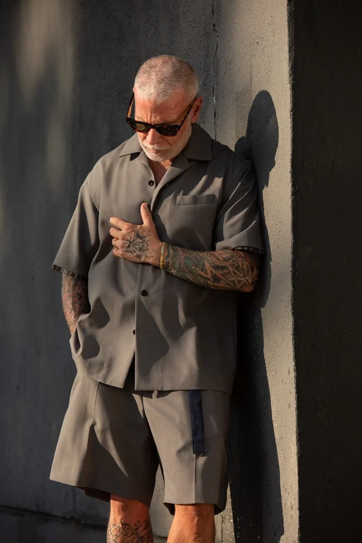 Older man with tattoos wearing grey short-sleeve shirt and matching shorts leaning against wall