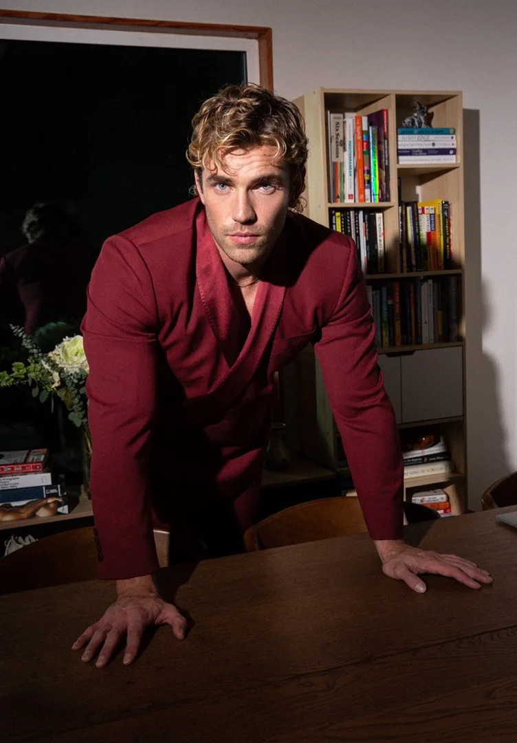 Adult male model with blonde hair in a red suit poses indoors with a table, bookshelf, and flowers in a lifestyle editorial shot.