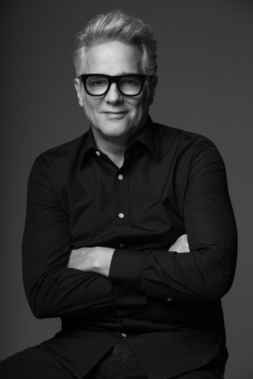 Monochrome portrait of an older man with glasses and grey hair wearing a black button-down shirt with arms crossed.