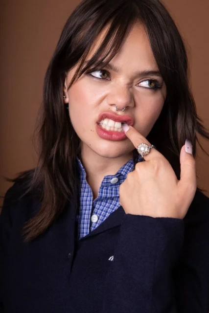Portrait of a young woman with dark hair and fair skin wearing a blue shirt, sweater, and ring, making a funny face indoors.