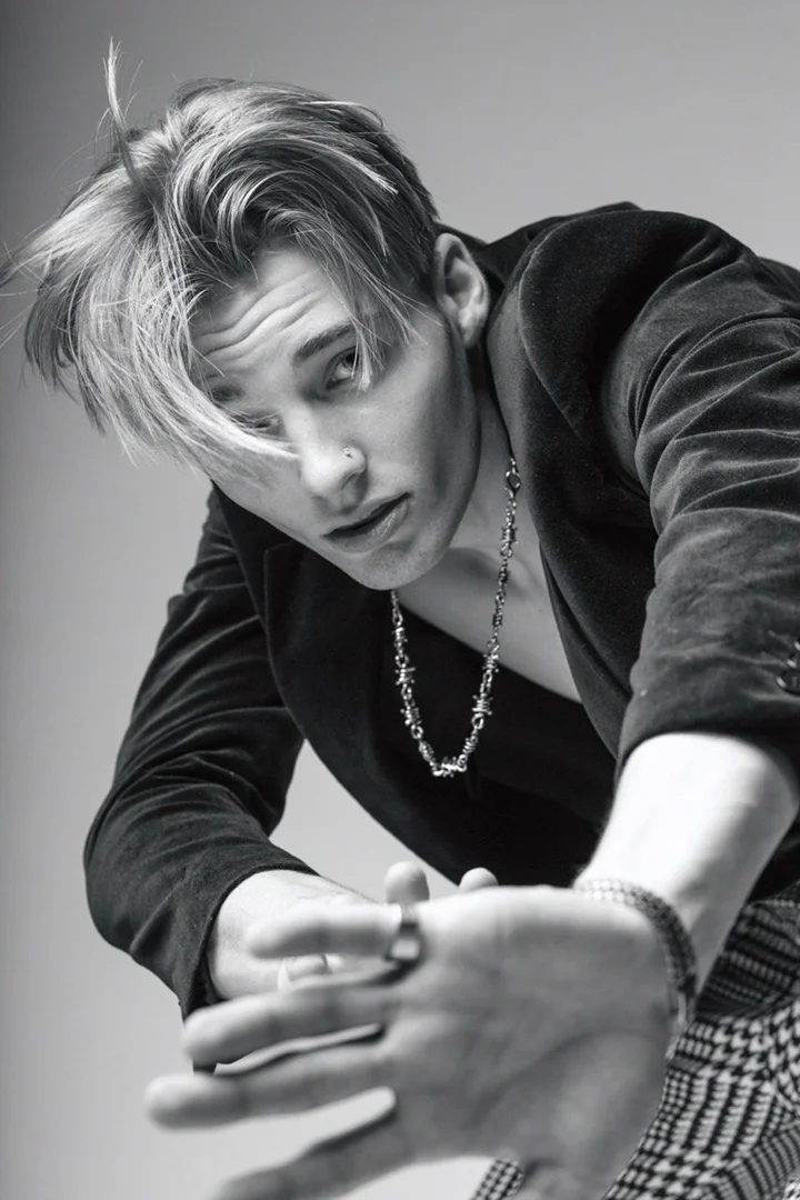 Monochrome portrait of a male model wearing a black velvet jacket and a silver necklace against a plain studio backdrop.
