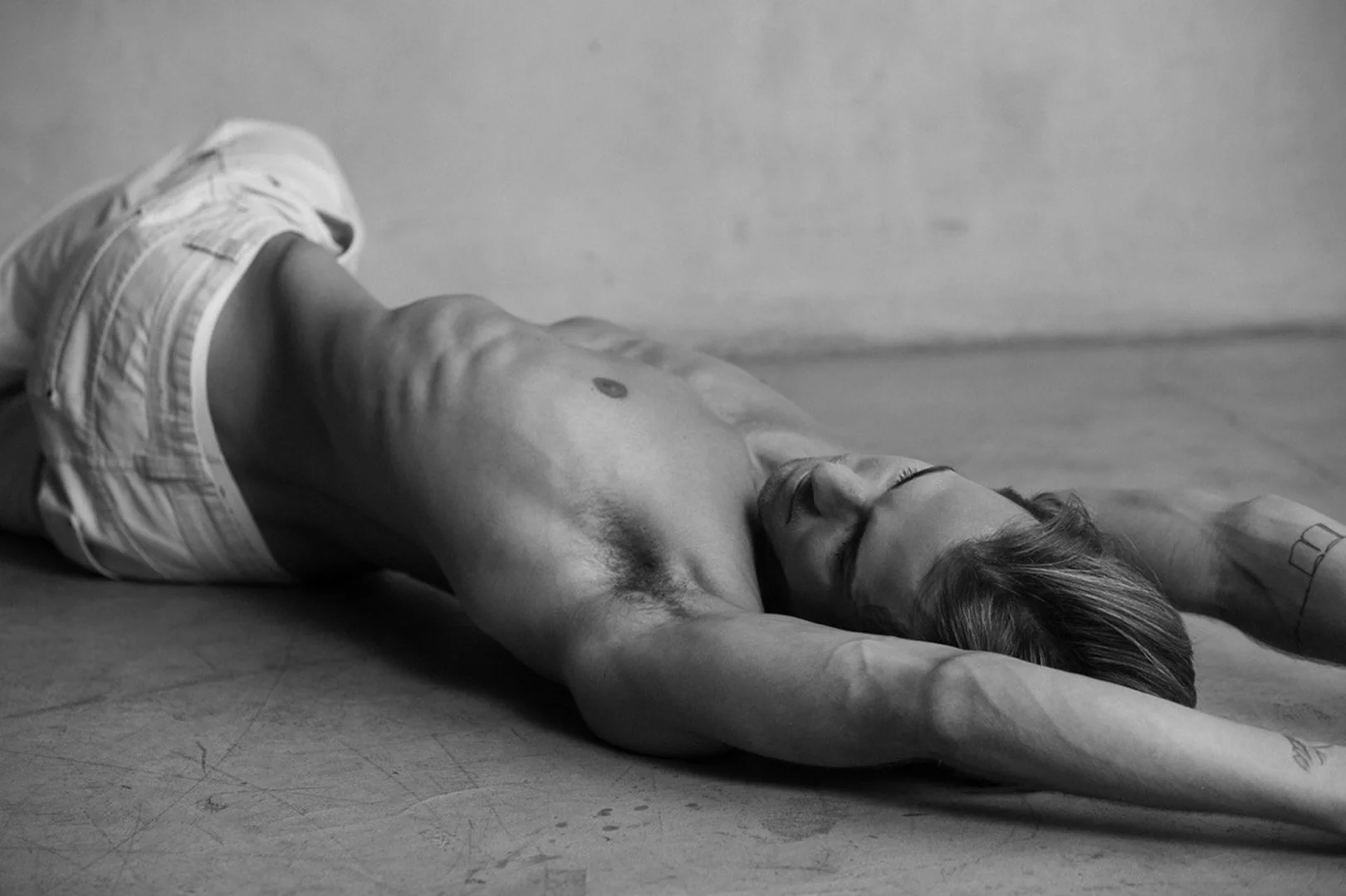 Black and white shot of an adult male with a muscular build, lying on a concrete floor in white shorts with an industrial background.