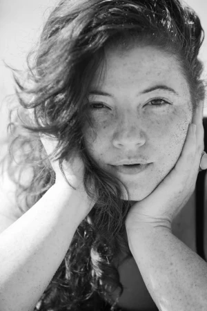 Close up monochrome portrait of a woman resting her face in her hands with dark curly hair and freckles.
