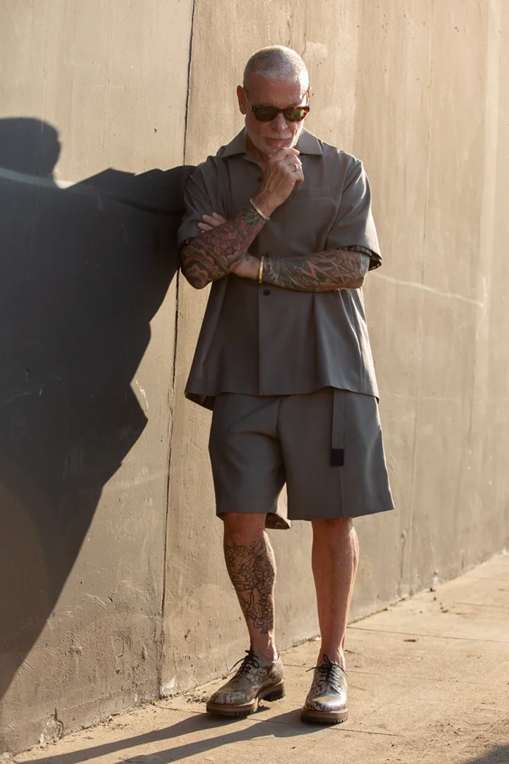 Mature man with tattoos in short sleeve button down and shorts leaning against a wall and posing in sunlight with sunglasses.