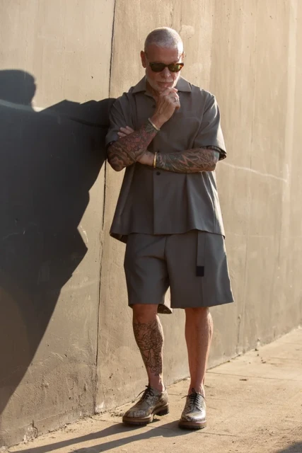 Mature man with tattoos in short sleeve button down and shorts leaning against a wall and posing in sunlight with sunglasses.
