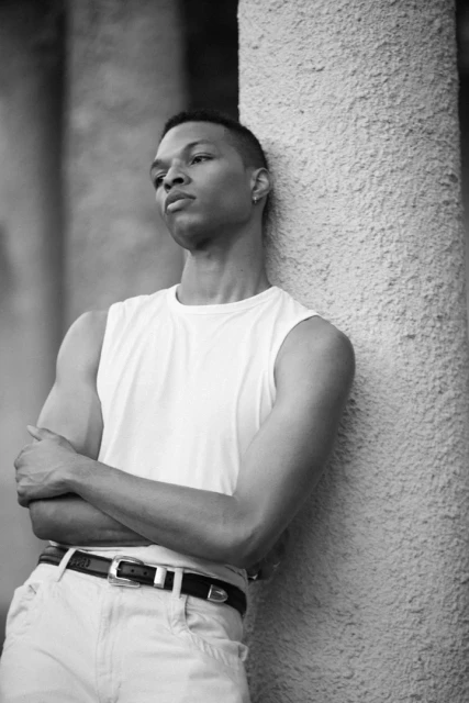 Black and white portrait of a young man with arms crossed, leaning against a textured stone column in an outdoor setting.