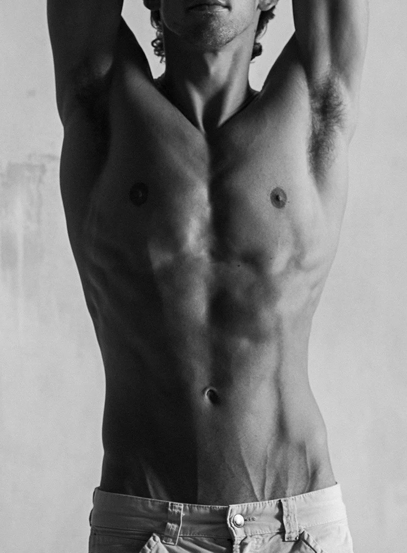 Black and white studio portrait of a muscular adult male torso, arms raised, wearing jeans against a neutral background.
