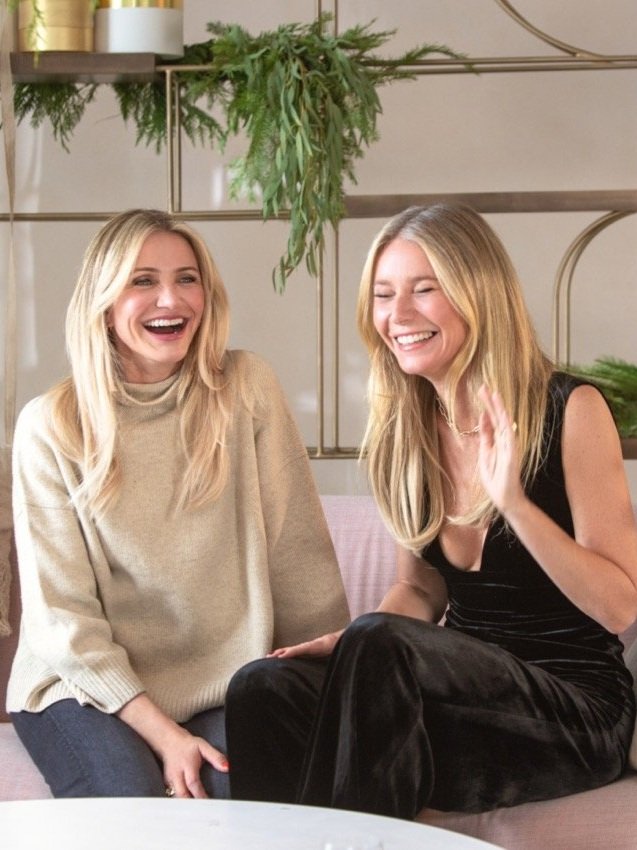 Two adult women are sitting indoors laughing together, one in a beige sweater and jeans, the other in a black velvet top and pants.