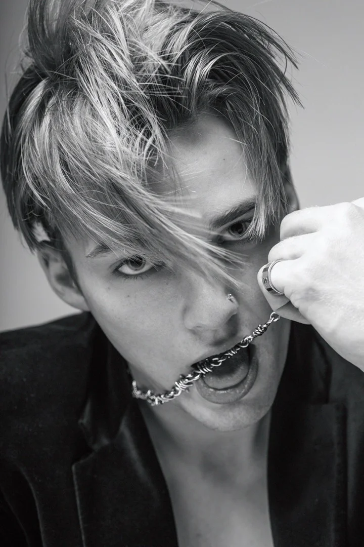 Black-and-white portrait of a young adult with blonde-streaked hair, wearing a jacket, holding a silver chain in the mouth.