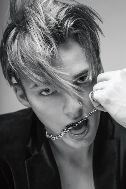 Black-and-white portrait of a young adult with blonde-streaked hair, wearing a jacket, holding a silver chain in the mouth.