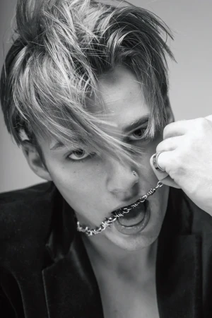 Black-and-white portrait of a young adult with blonde-streaked hair, wearing a jacket, holding a silver chain in the mouth.