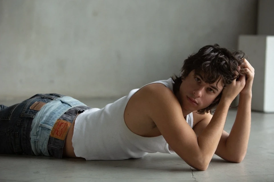 Young man lying down with hands behind his head wearing a white tank top and jeans in a grey interior with soft natural light.