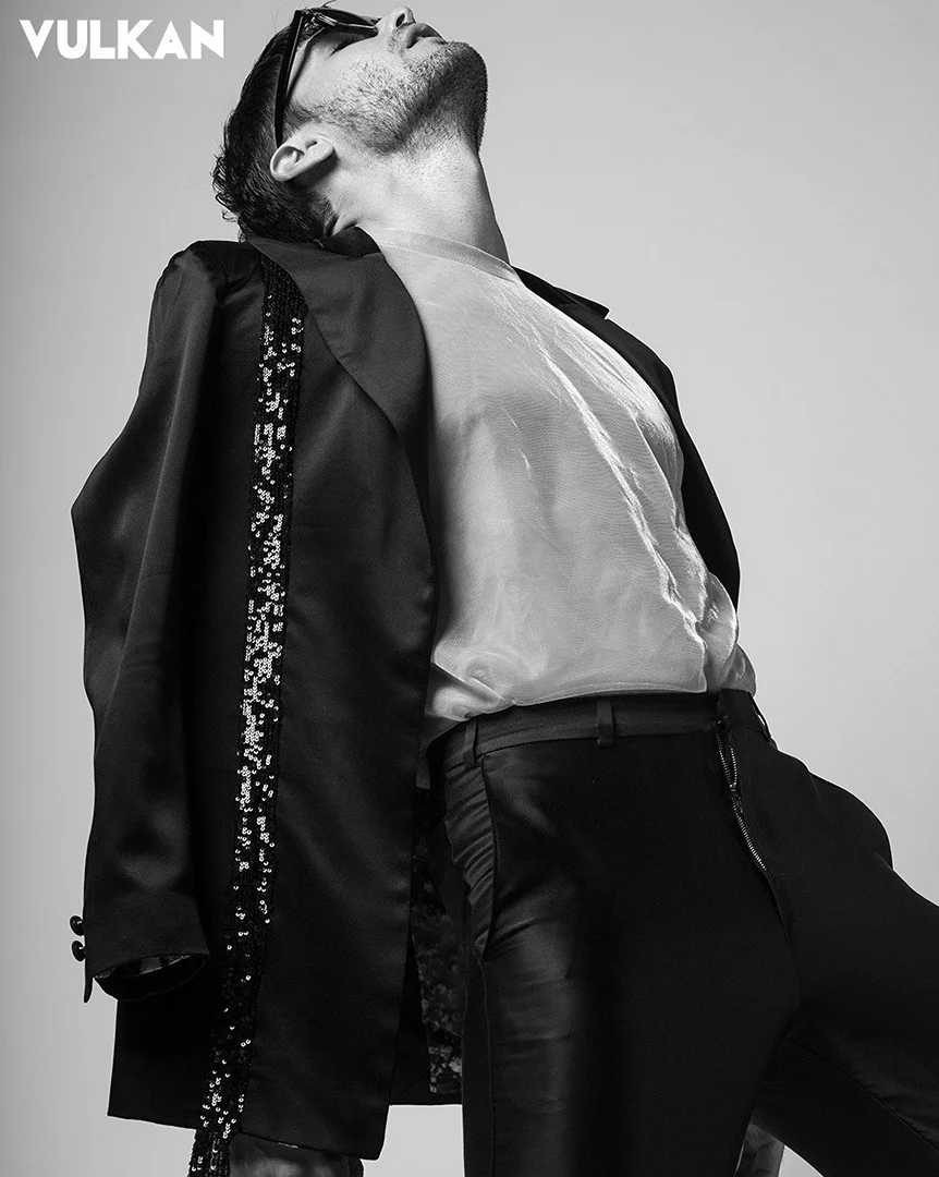 Black and white studio shot of a man wearing sunglasses, a sheer t-shirt, jacket, and pants, posing with his head tilted back.